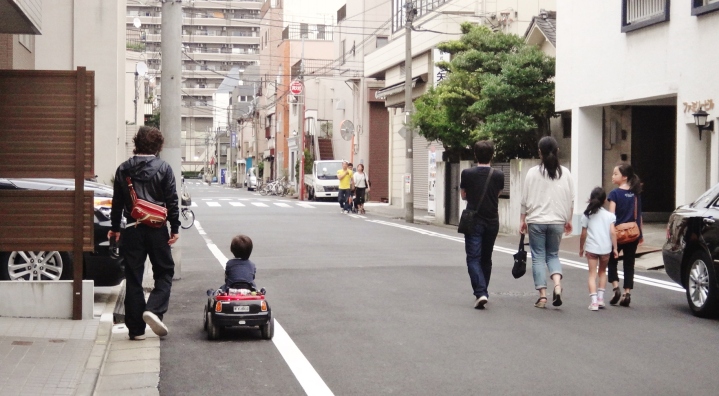 A child rides a miniature car on a narrow Tokyo street, next to a family of four.